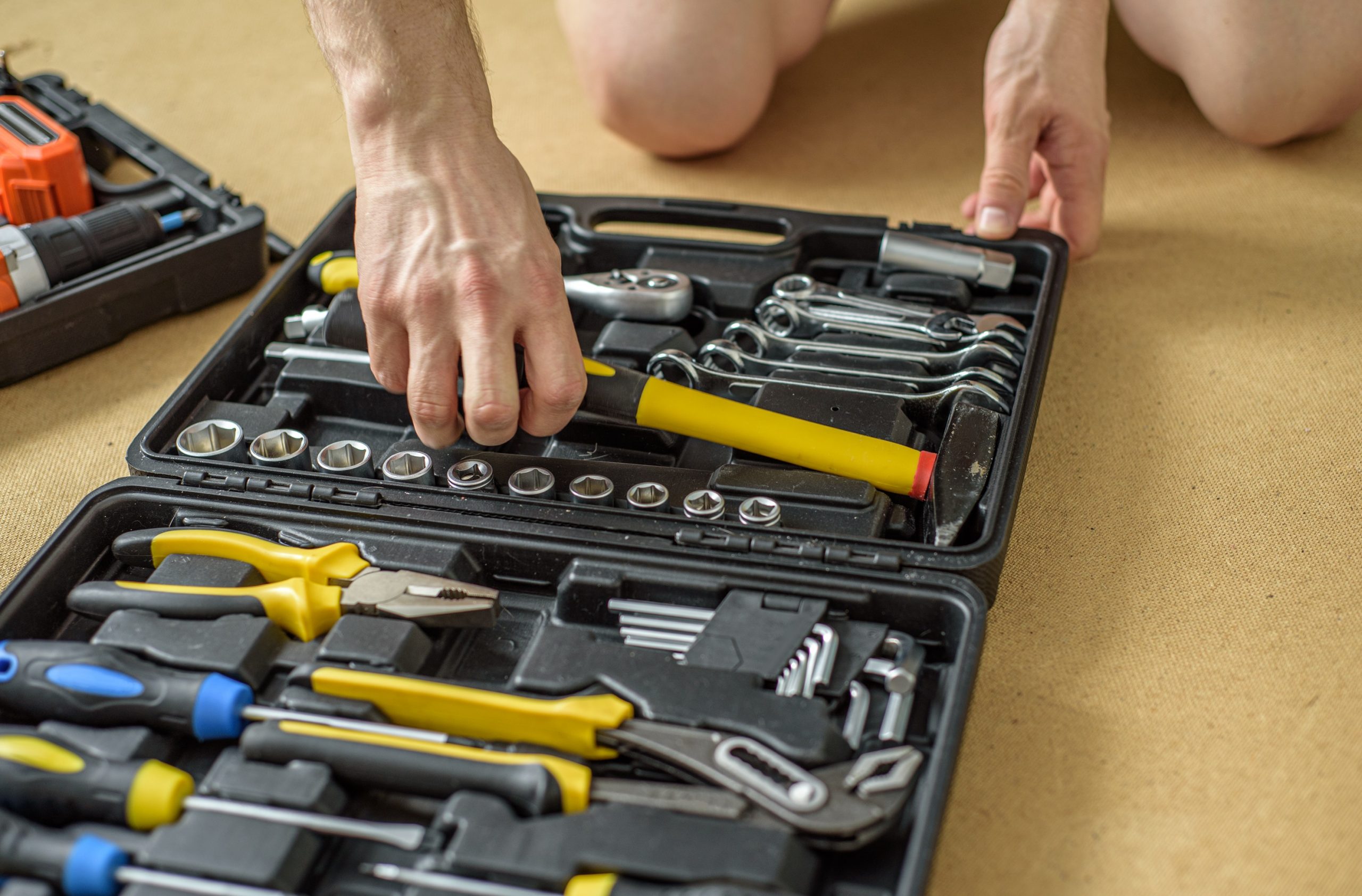 Na imagem, um homem está agachado, enquanto organiza uma caixa de ferramentas. A caixa está aberta apoiada na chão, com várias ferramentas dentro dela.