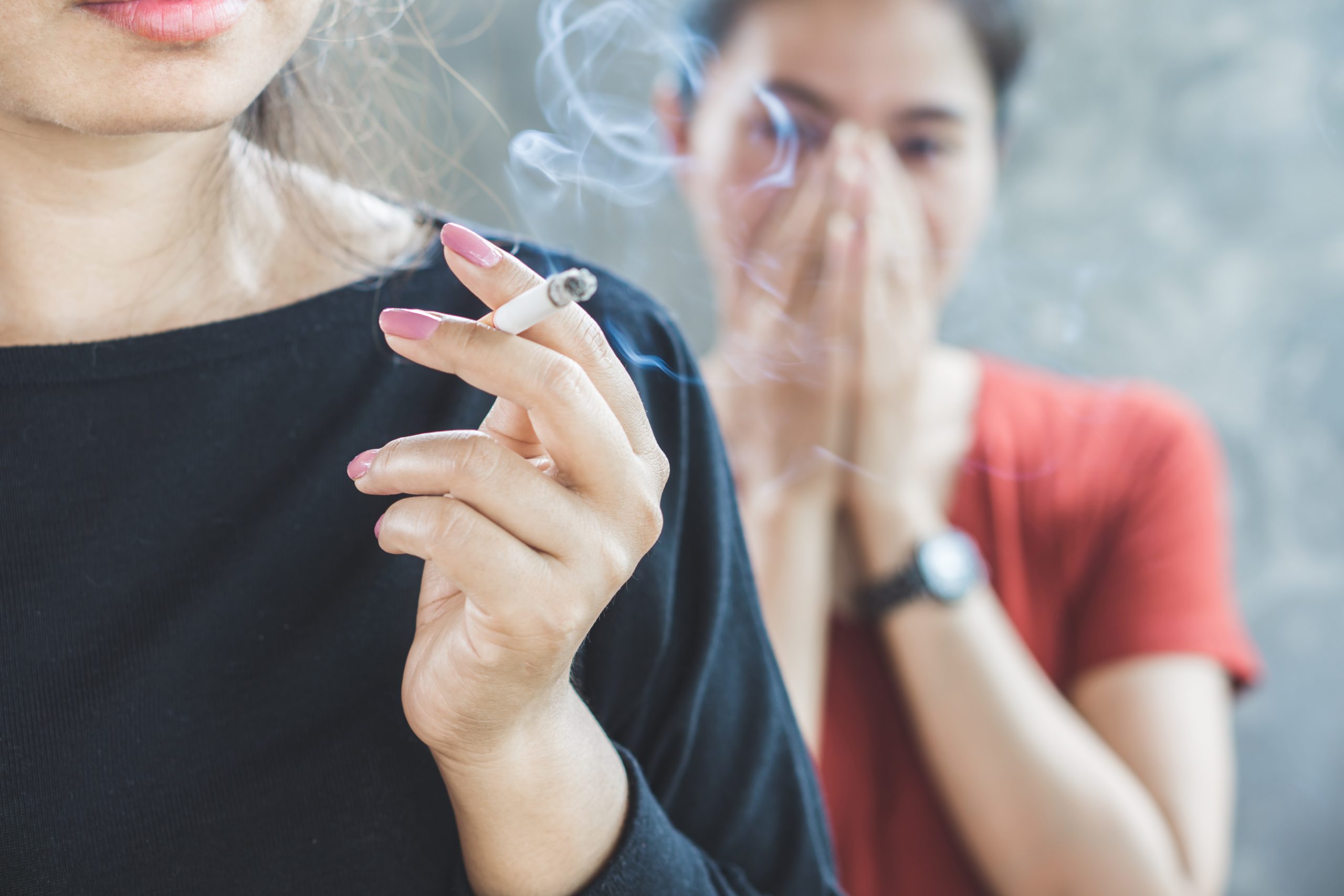 Na imagem uma mulher com blusa preta está segurando um cigarro. Ao fundo, uma mulher de blusa vermelha está tampando o nariz, por conta do cheiro de cigarro