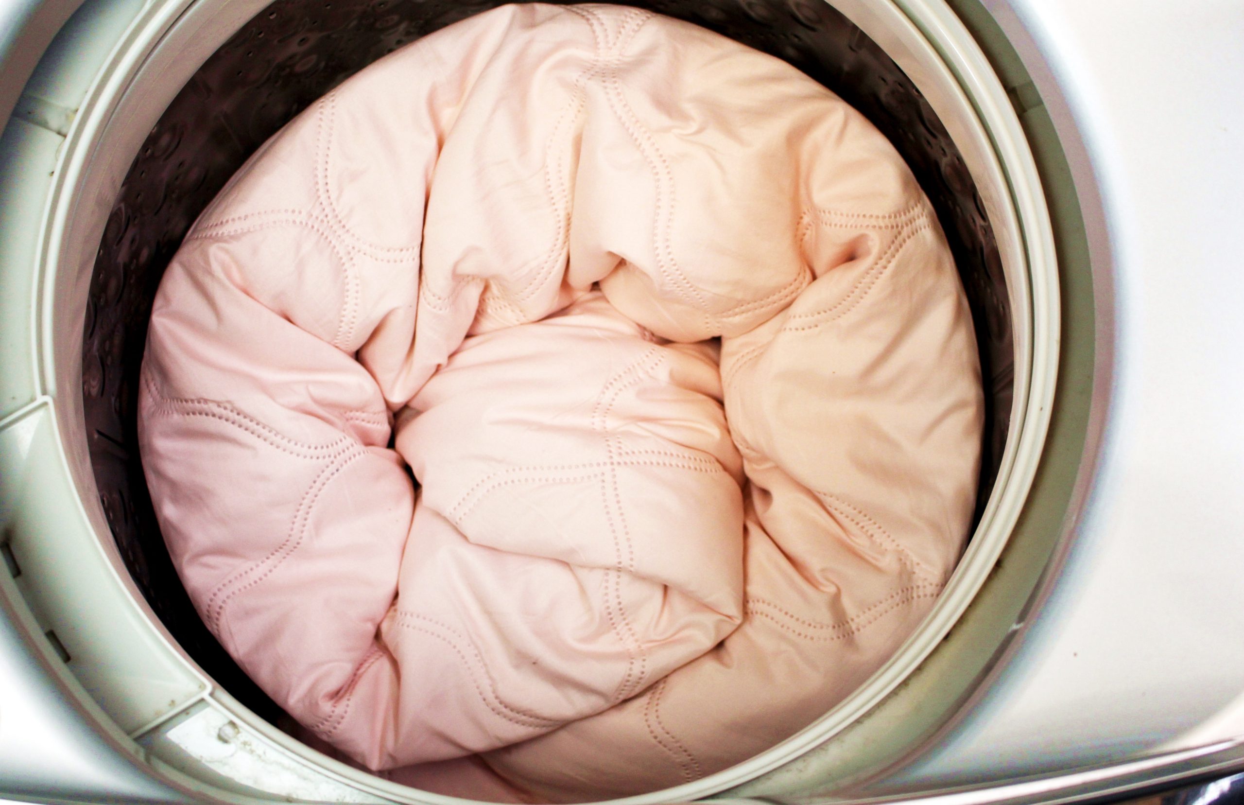 Na imagem é possível ver uma edredom na cor rosa dentro de uma maquina de lavar
