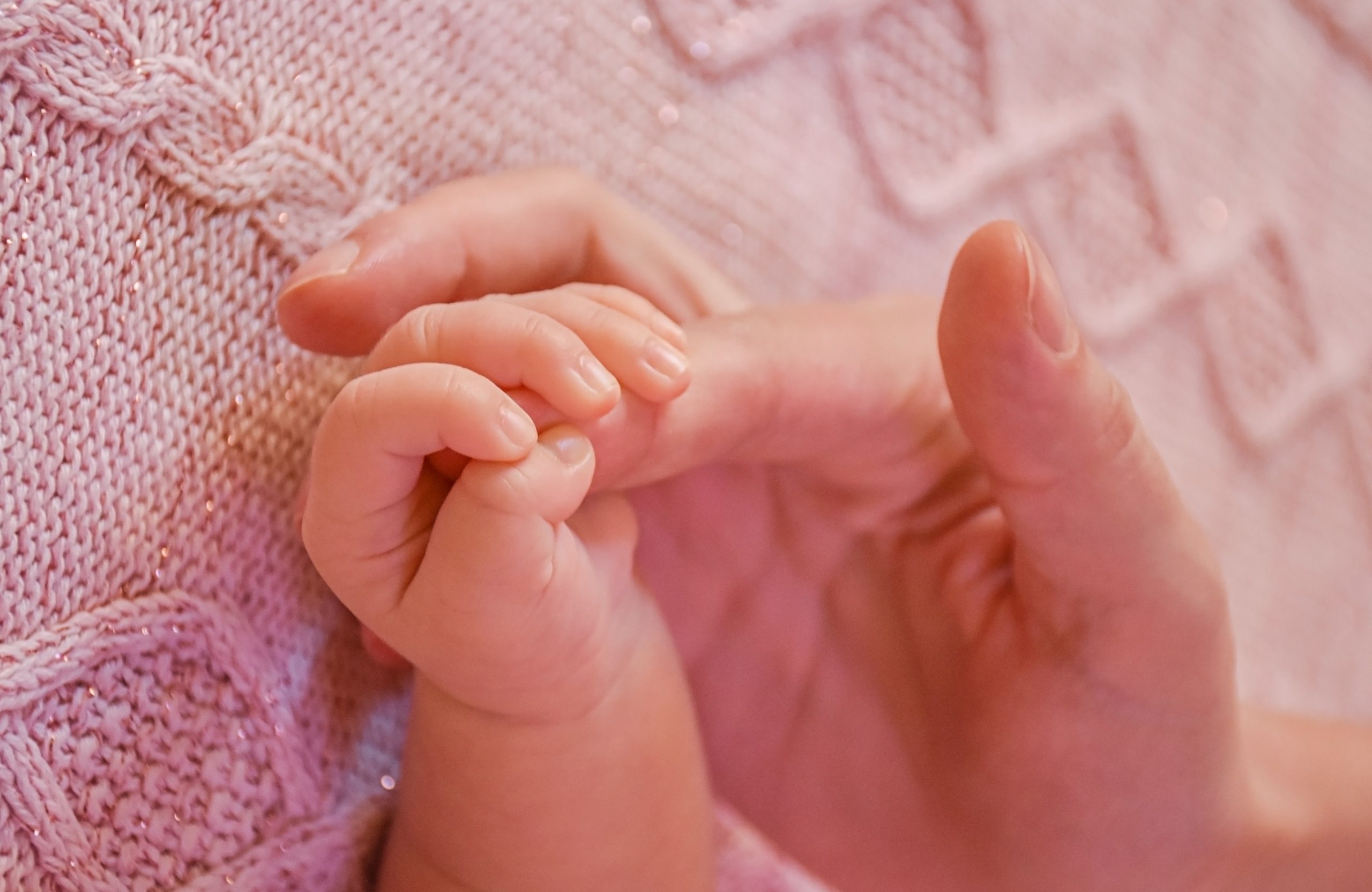 Visão parcial do braço esquerdo de um bebê segurando o dedo indicador da mão de um adulto. Ao fundo observa-se uma manta rosa
