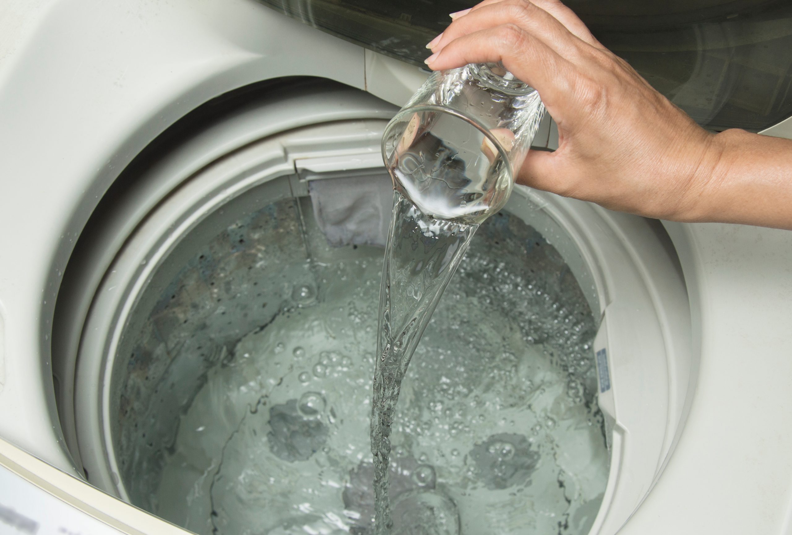Na imagem, é possível visualizar uma maquina de lavar com sua tampa levantada e dentro dela é possível ver água até o nível médio. Também é possivel ver uma pessoa adicionando com o auxilio de um copo um liquido dento da máquina