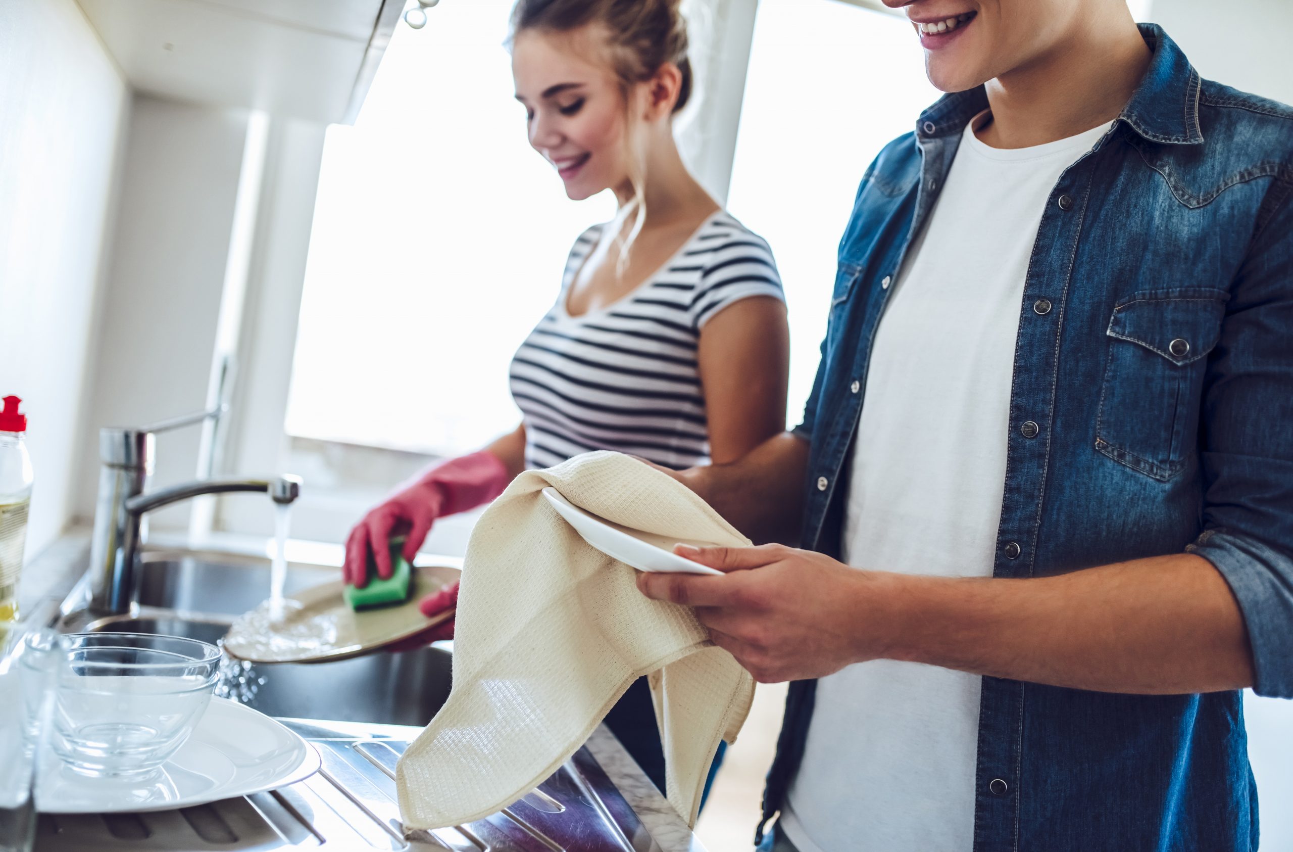 Na imagem, um mulher está lavando um prato na cor branca com o auxilio de uma esponja verde, ela usa uma luva na cor rosa. Ao seu lado está um homem secando um prato com o auxílio de um pano de prato.