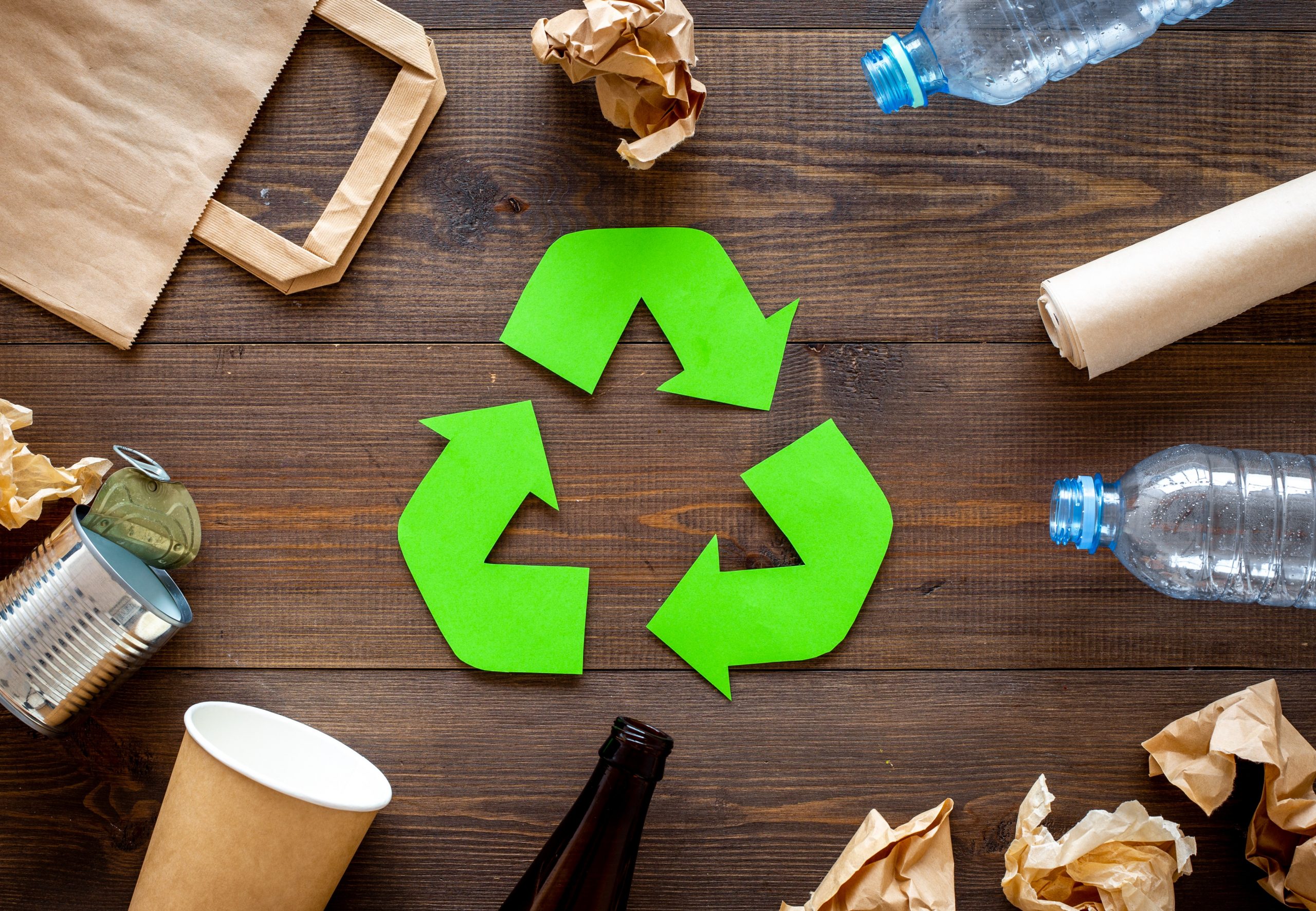 Três setas verdes, formando o símbolo de reciclável sobre uma superfície de madeira rodeada de garrafas de plástico, de vidro, lata de alumínio e papel.