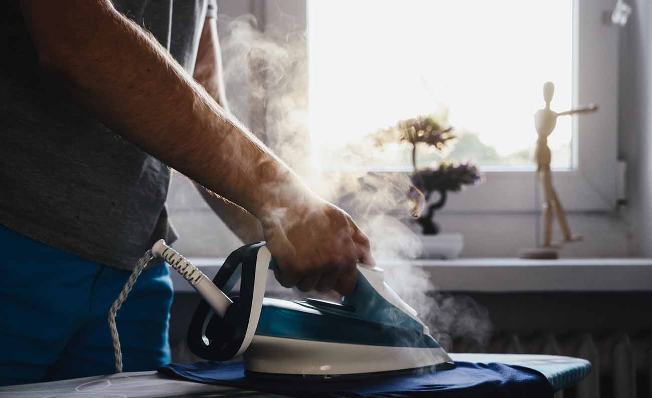 Imagem cortada de um homem diante de uma tábua de passar roupa, segurando um ferro de passar roupa liberando vapor