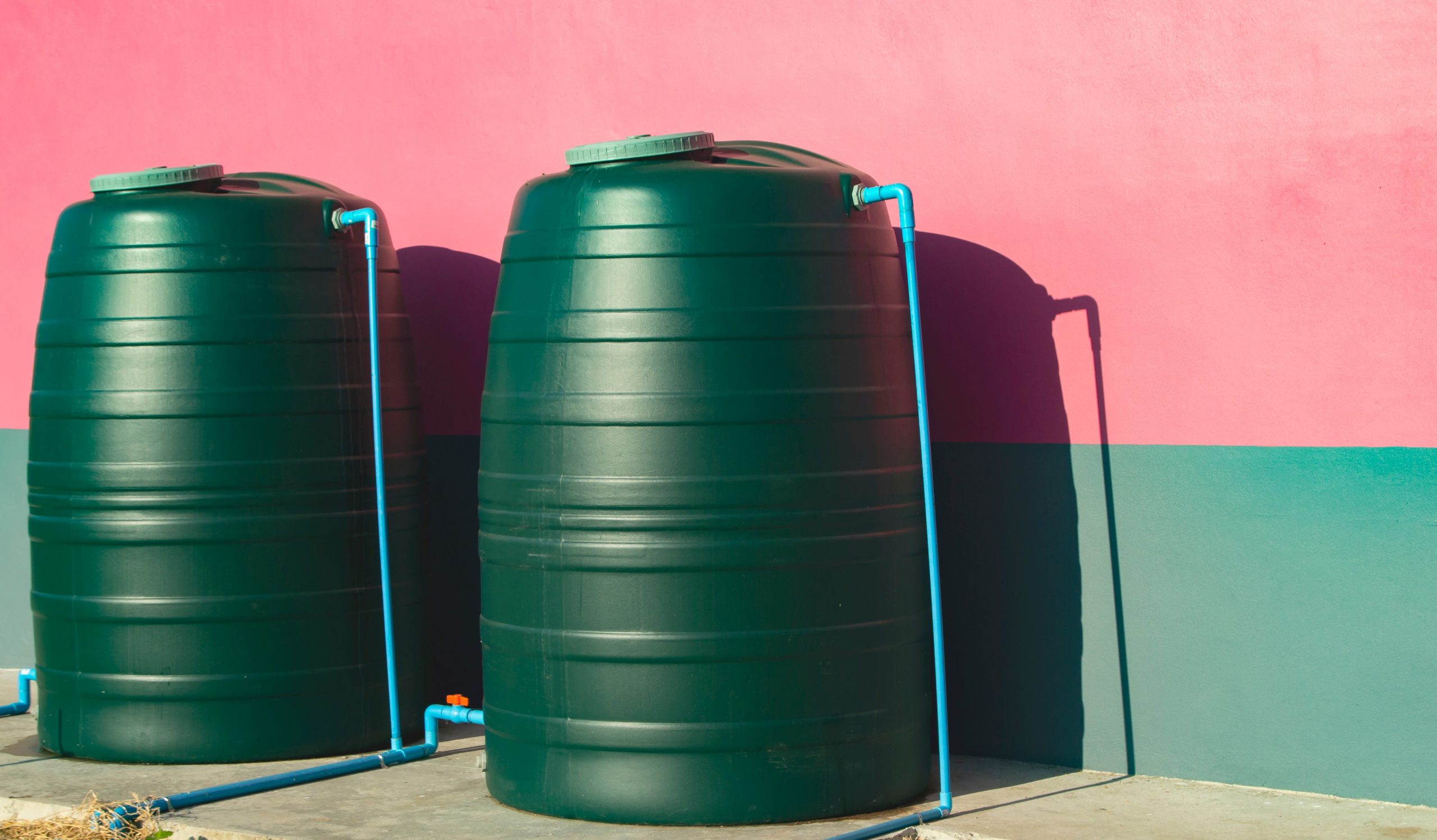 Duas cisternas de plástico da cor verde, conectada por canos azuis. As cisternas estão sobre uma caçada de cimento e a cor da parede logo atrás é metade verde e metade vermelha
