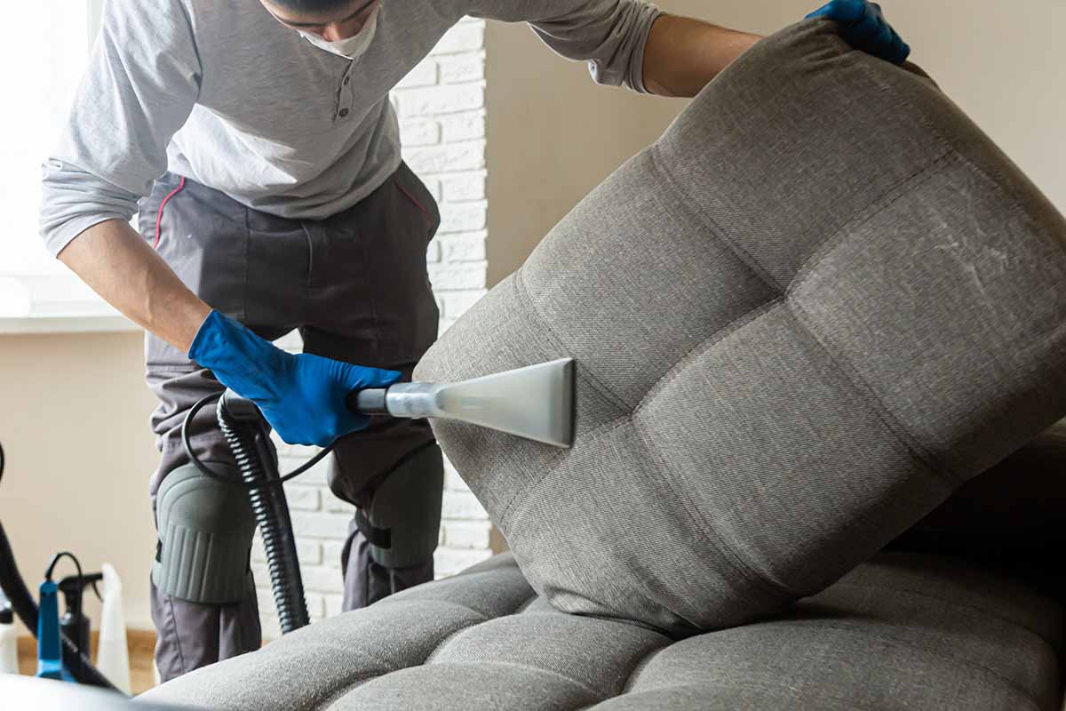 Imagem cortada de um homem limpando um sofá na cor cinza, como auxilio de um aspirador para retirar a sujeira.