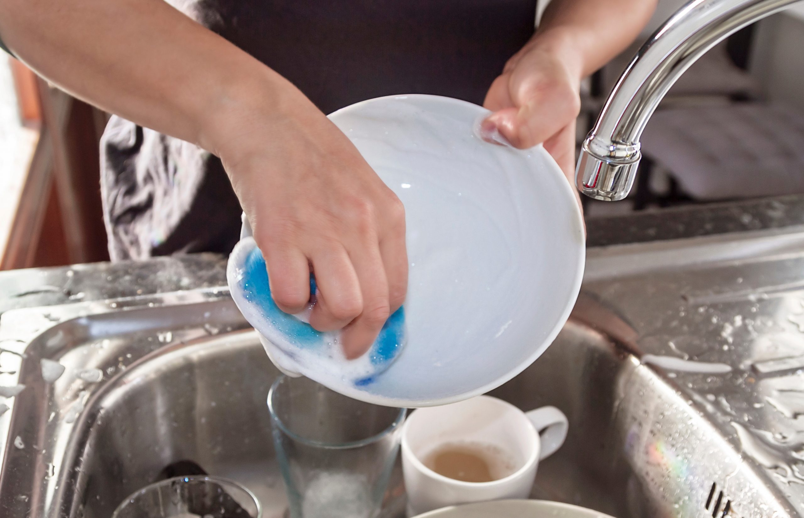 Pessoa com os braços estendidos sobre uma pia inox com poucas peças para lavar. Nas mãos a pessoa ensaboa um prato branco. A torneira está fechada