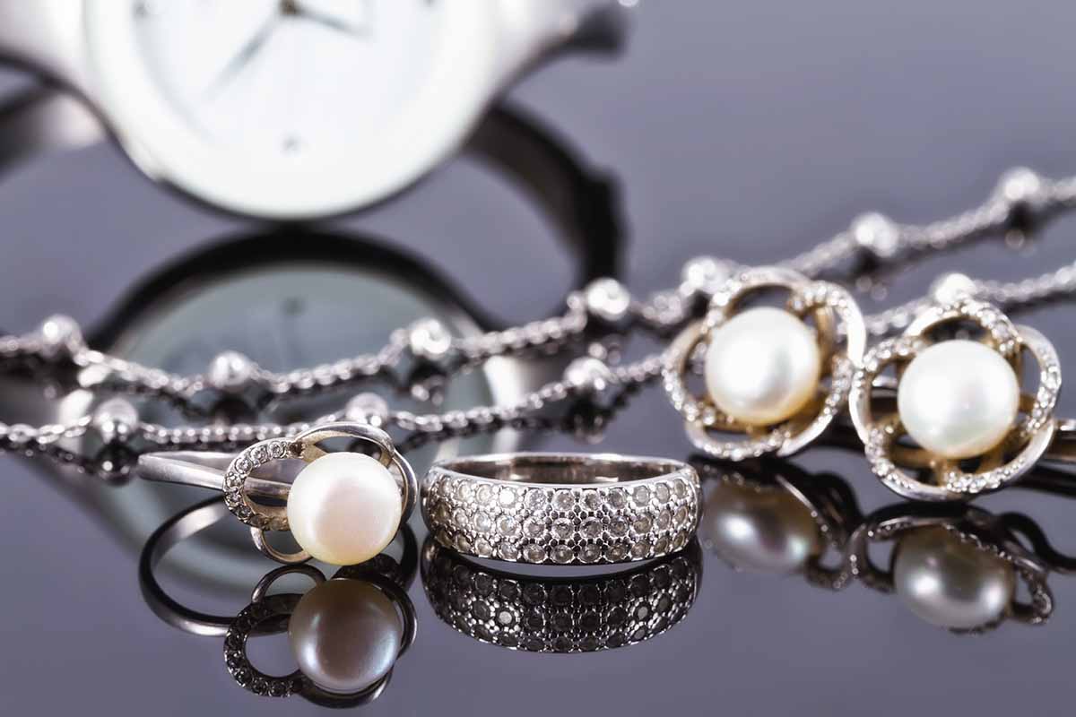 magem cortada de um balcão com jóias expostas. É possível encontrar três anéis de pérolas com adornos em prata, um anel prata cravejado com brilhos, um colar prata e ao fundo um relógio também prata.