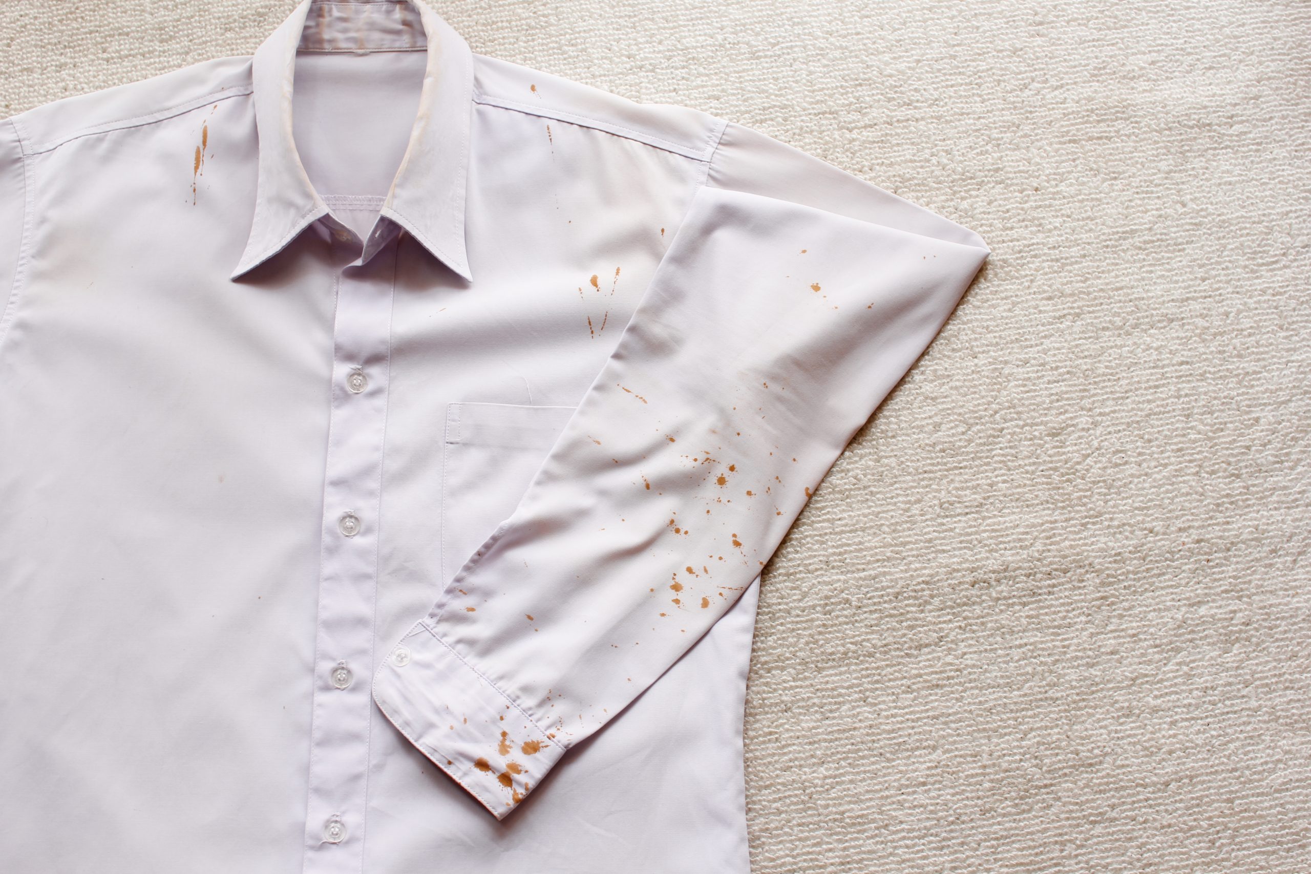 Camisa social clara e de botões, estendida sobre uma superfície reta com o braço esquerdo dobrado sobre o corpo da camisa, salpicado por diversas manchas de ferrugem.