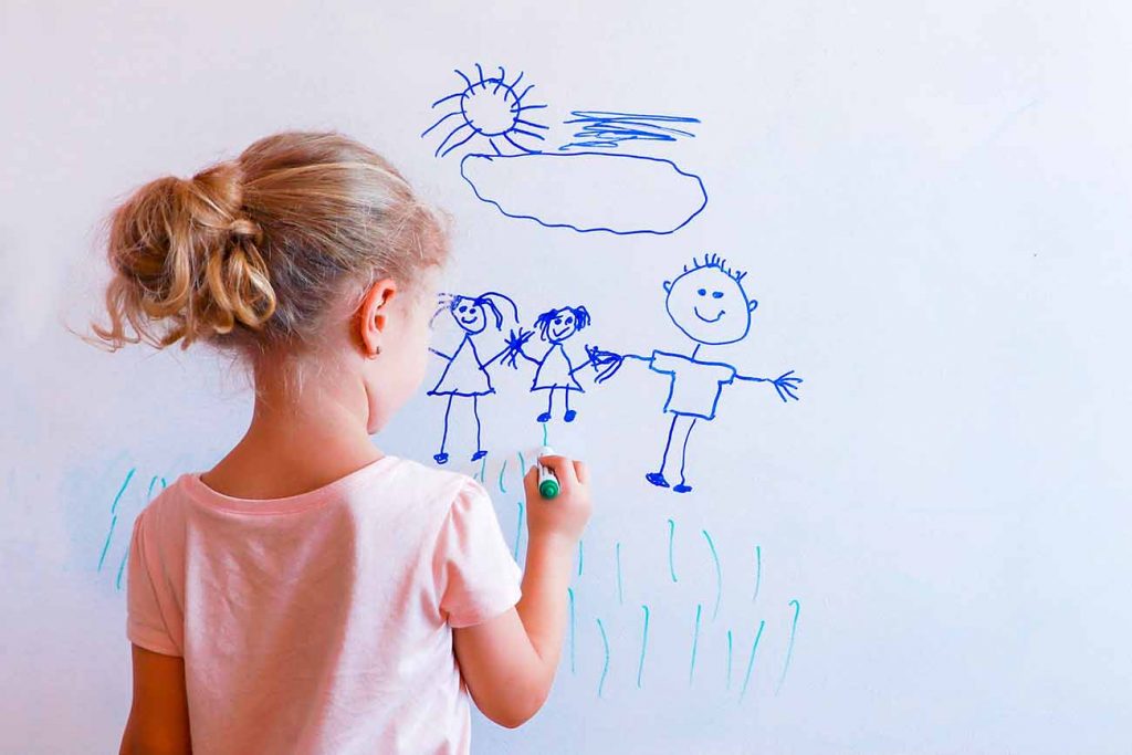 Criança de costas diante de uma parede branca com uma canetinha nas mãos, desenhando na parede