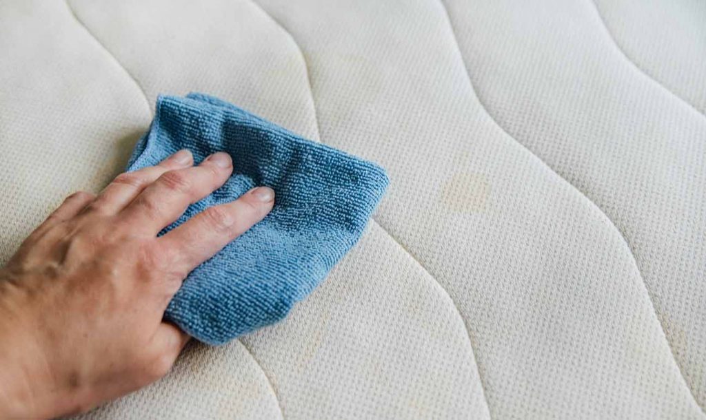 magem cortada de mão com um pano de limpeza azul limpando a superfície encardida de um colchão. 