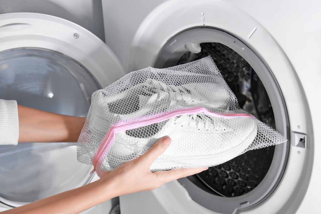 Imagem cortada dos braços de uma pessoa segurando um par de tênis dentro de saco de lavagem de roupas, diante de uma máquina de lavar roupas, com abertura frontal, aberta